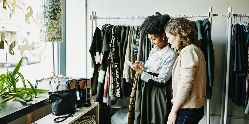 Smiling woman showing shop owner photos on smartphone while shopping in boutique shop